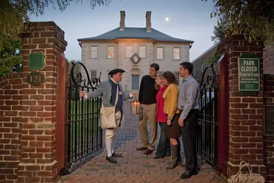A man in historical costume holding a lantern welcomes four casually dressed adults at the entrance gate of a brick building with lit windows at dusk. Signs and iron gates frame the entrance.