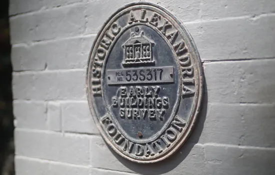 A round metal plaque on a white brick wall reads Historic Alexandria Foundation, Early Buildings Survey, Reg. No. 535317 with a small building illustration at the top.