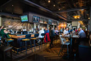 A lively sports bar with patrons sitting at the bar and tables, watching multiple TVs displaying sports. The decor features a football field mural and modern lighting, creating a vibrant, social atmosphere.