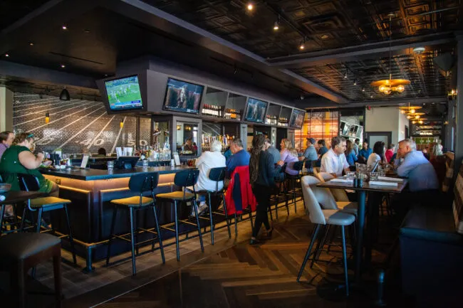 A lively sports bar with patrons sitting at the bar and tables, watching multiple TVs displaying sports. The decor features a football field mural and modern lighting, creating a vibrant, social atmosphere.