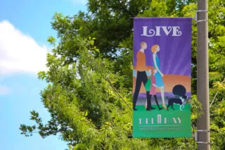 A street banner shows an illustrated family with a dog, the word LIVE above them, and text below that reads Del Ray: Where Main Street Still Exists. Green trees and blue sky are in the background.