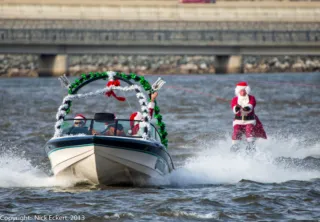 A person dressed as Santa Claus is water skiing behind a Holidays-themed boat decorated with green garland and ornaments. Two people in Santa hats ride in the boat on a body of water near a bridge.