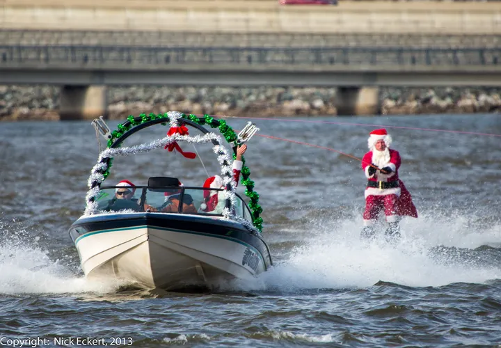 A person dressed as Santa Claus is water skiing behind a Holidays-themed boat decorated with green garland and ornaments. Two people in Santa hats ride in the boat on a body of water near a bridge.