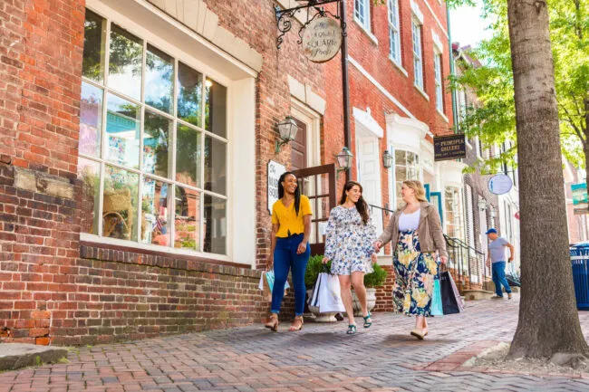 Three women walk down a brick sidewalk, carrying shopping bags and smiling, in front of shops with large windows and hanging signs on a sunny day. Trees line the street, and a man is seen in the background.