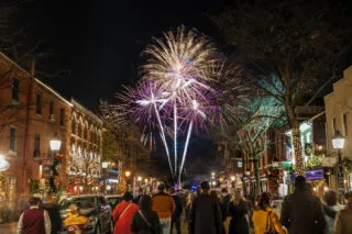 A crowd gathers on a festive city street at night during the holidays, watching colorful fireworks burst in the sky above brick buildings decorated with lights.