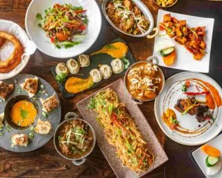 An overhead view of a table filled with a variety of vibrant Indian and Asian dishes, including noodles, curries, grilled meats, dumplings, naan, and colorful garnishes arranged on different plates and bowls.