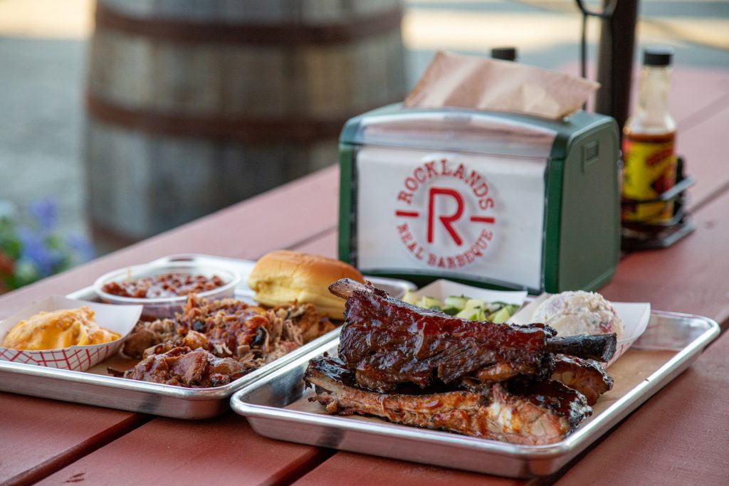 Trays of barbecue food, including ribs, pulled pork, beans, mac and cheese, coleslaw, a sandwich, and pickles, sit on a picnic table next to a napkin holder labeled Rocklands Real Barbecue.
