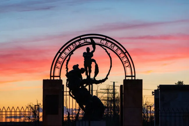 Silhouette of the Freedman’s Cemetery monument at sunset, with vibrant pink and orange clouds in the sky. The arched sign and statue are framed by fencing and pillars, with the word Cemetery visible.