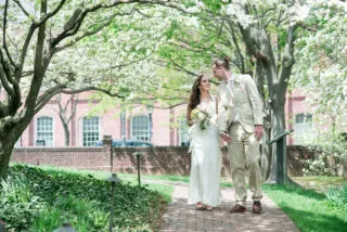A bride and groom walk hand in hand down a brick path under blooming trees, smiling at each other. The bride wears a white dress and holds a bouquet; the groom wears a beige suit. A brick building is visible in the background.