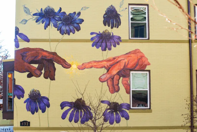 A mural on a yellow building depicts two large hands reaching toward each other with fingers almost touching, surrounded by purple flowers. A burst of light appears between the fingertips. Two windows are incorporated into the artwork.