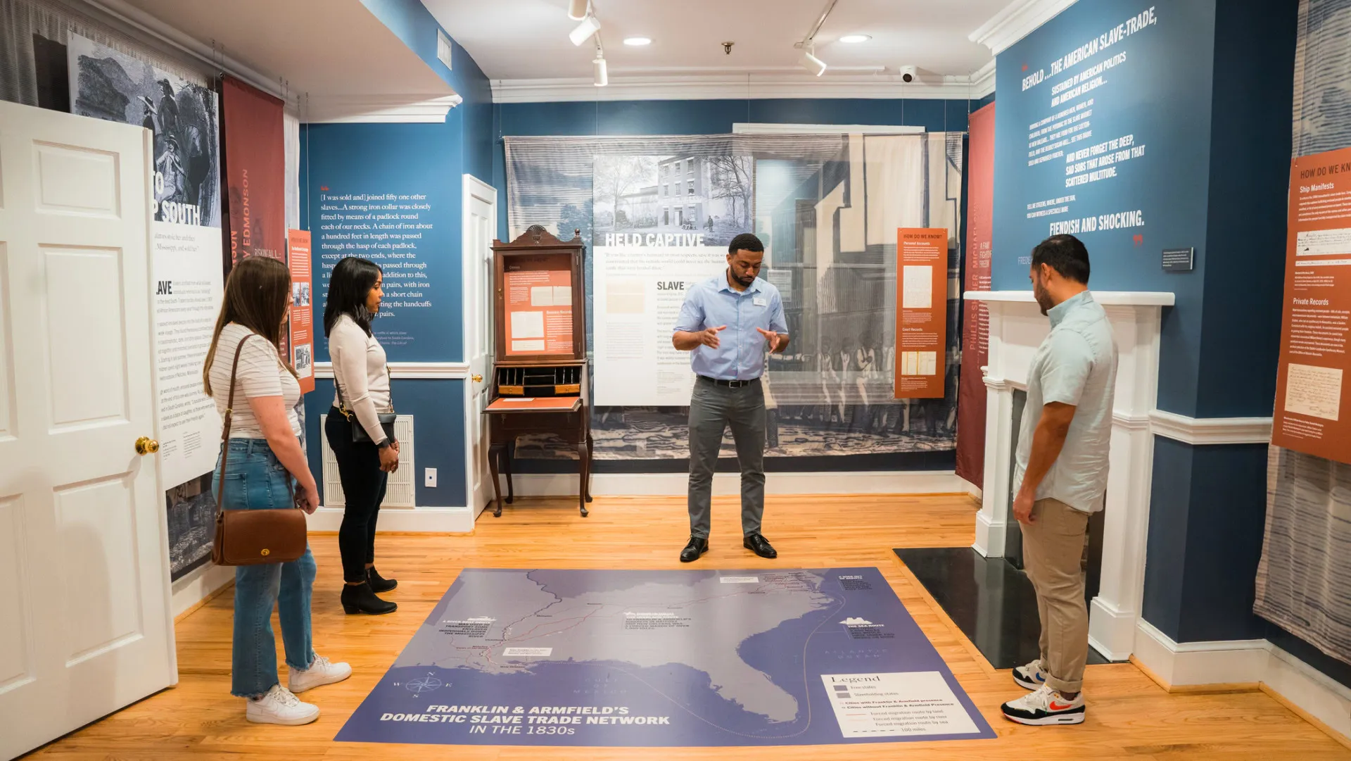 Four people stand in a museum exhibit room with informational signs on the walls and a map on the floor, listening to a guide explain the history of slavery in America.