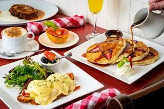 A brunch table set with pancakes topped with strawberries and syrup, eggs Benedict with salad, grilled grapefruit, a cappuccino, a glass of orange juice, and a red checkered napkin.