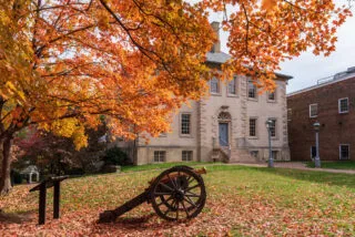 A historic stone building stands behind a cannon on a grassy lawn, surrounded by vibrant fall foliage and fallen leaves in shades of orange.