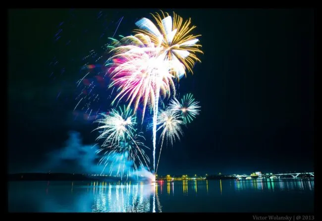 Colorful fireworks burst over a calm body of water at night; their reflections shimmer on the surface, with city lights and a bridge visible in the distance under a dark sky.