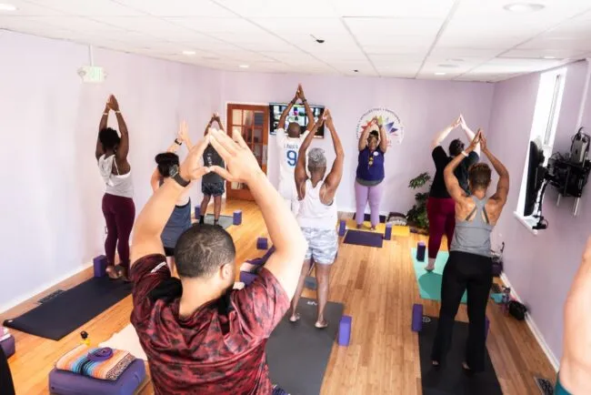 A diverse group of people practice yoga together in a brightly lit studio, standing on mats with arms raised overhead in a pose, facing the instructor at the front of the room.