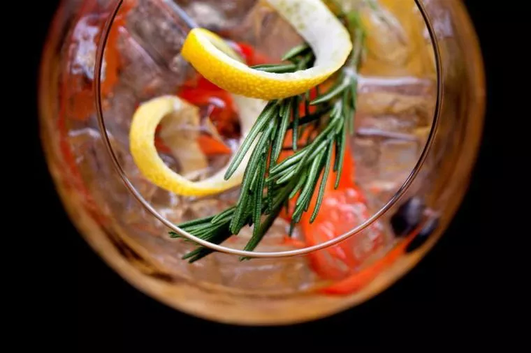 A close-up, top-down view of a cocktail with ice, a twist of lemon peel, a sprig of rosemary, and red berries in a clear glass against a black background.