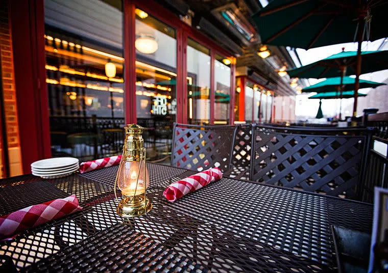 A close-up of a metal outdoor dining table set with neatly rolled red-and-white napkins, plates, and a lit lantern. The background features patio umbrellas and a building with large windows.