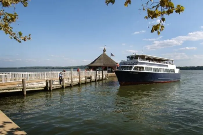 A white and blue sightseeing boat is docked at a wooden pier on a lake, with a small pavilion, a few people on the dock, and trees in the background under a blue sky with scattered clouds.