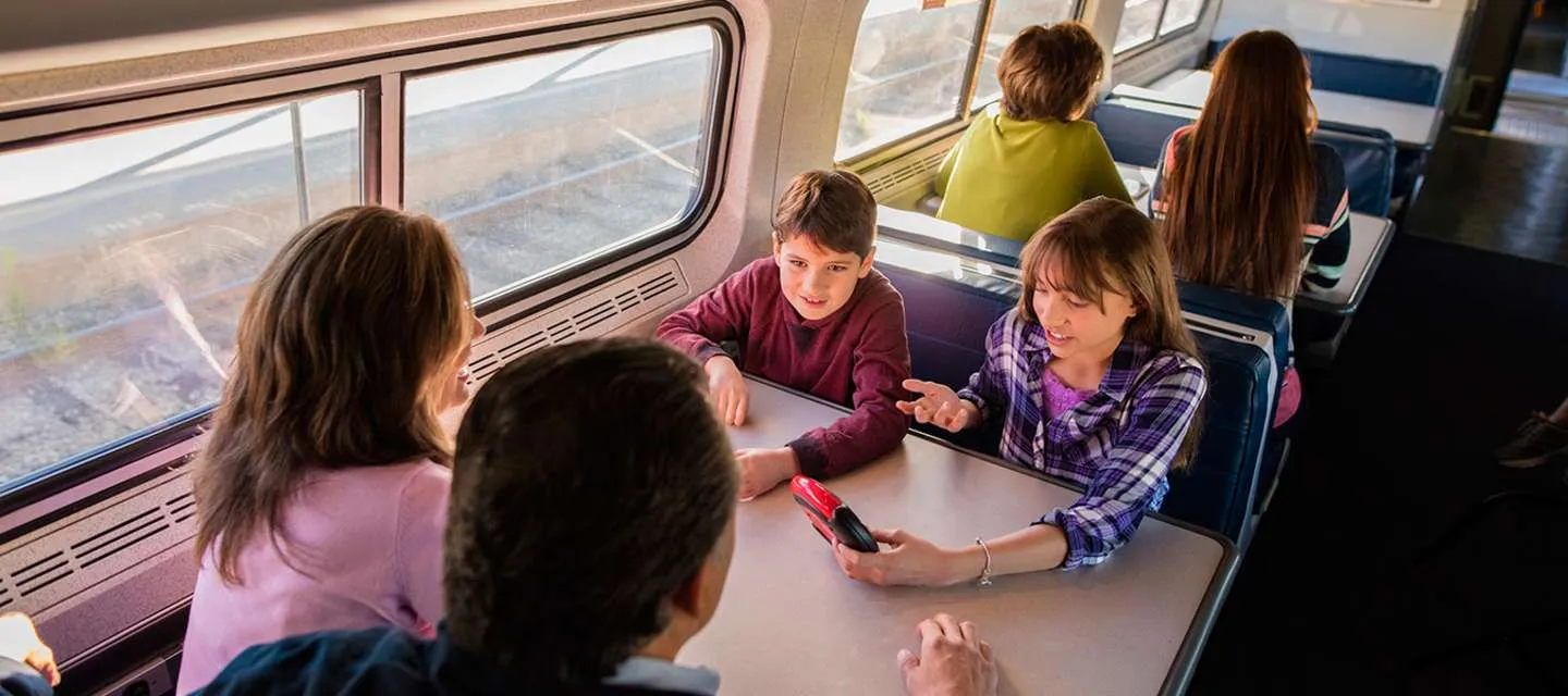 A family of four sits at a table on a train, talking and smiling. Two children face the camera, with one holding a phone, while two adults sit across from them. Other passengers are visible in the background.