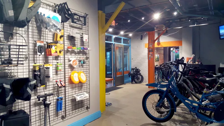 A bike shop interior with bikes lined up on the right and various cycling accessories displayed on a wall grid to the left; bright yellow and orange beams, and a glass door leading to another room are visible.