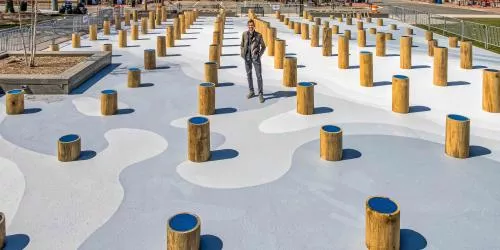 A person stands among many short wooden posts with blue tops, set in an outdoor area with abstract white and gray patterns on the ground.