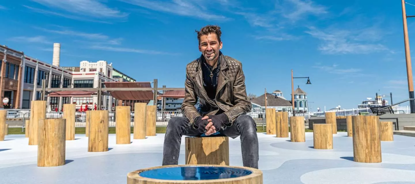 A man wearing a brown leather jacket and black jeans sits on a wooden post in an outdoor urban area surrounded by similar wooden posts, with buildings and a blue sky in the background.