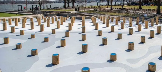 A playground features numerous short, cylindrical wooden posts spaced evenly across a flat, painted surface with wavy patterns. Trees, grass, a river, and a bridge appear in the background.