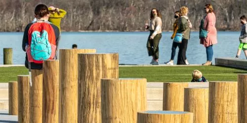 A person with a turquoise backpack sits on wooden posts by the water, while several people walk and stand near the shore in the background. Trees with no leaves are visible across the water.