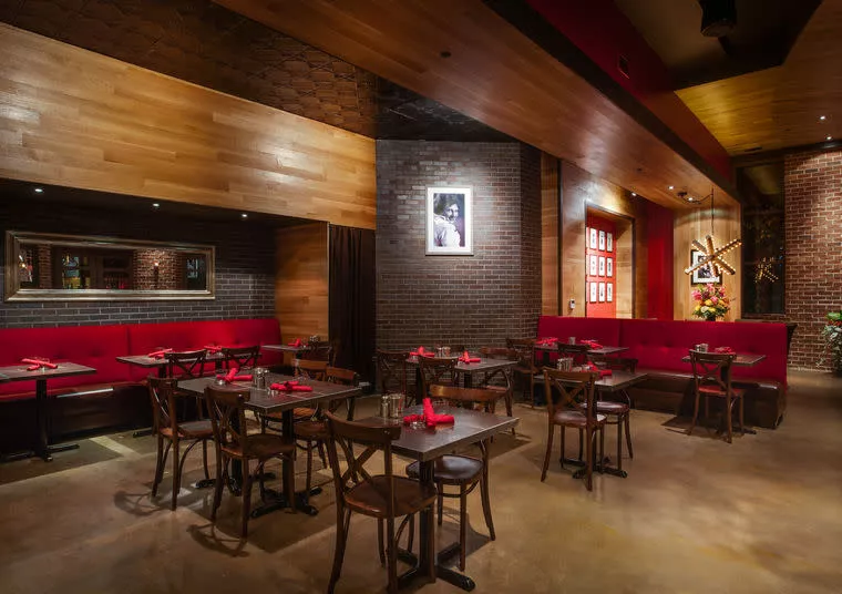 A modern restaurant interior with wooden ceilings, exposed brick walls, red upholstered booths, dark wood tables and chairs, framed photos, and dim, warm lighting. Tables are neatly set with red napkins.
