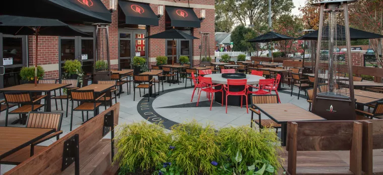 Outdoor restaurant patio with wooden tables and chairs, black umbrellas, red chairs around a central round table, potted plants, and a tall outdoor heater, all set against a brick building.