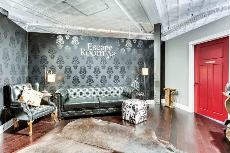 A stylish lounge with a black leather sofa, armchair, cowhide ottoman, and rug. The wall reads Escape Room in white letters. A red door and vintage decor complete the room’s elegant, cozy look.
