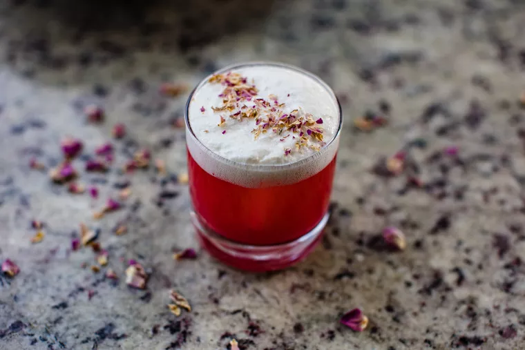 A vibrant red drink in a short glass sits on a speckled countertop, topped with a thick layer of foam and sprinkled with dried rose petals. Scattered petals surround the glass.