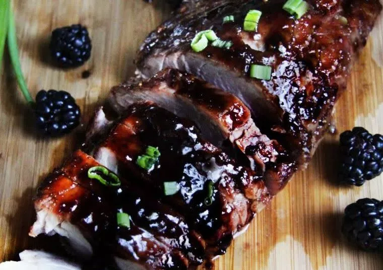 Sliced glazed pork tenderloin on a wooden board, garnished with chopped green onions and surrounded by fresh blackberries.