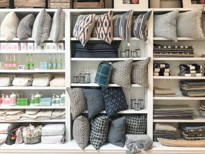 Shelves filled with decorative pillows in blue, grey, and neutral patterns, alongside neatly arranged towels, soaps, and storage baskets in a bright, organized home goods store.
