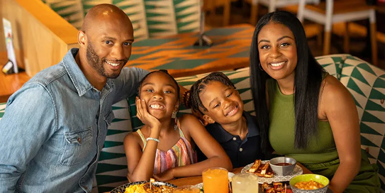 A smiling family of four sits together at a restaurant table with food and drinks, posing for a cheerful photo. The two children sit between the adults, all appearing happy and relaxed.