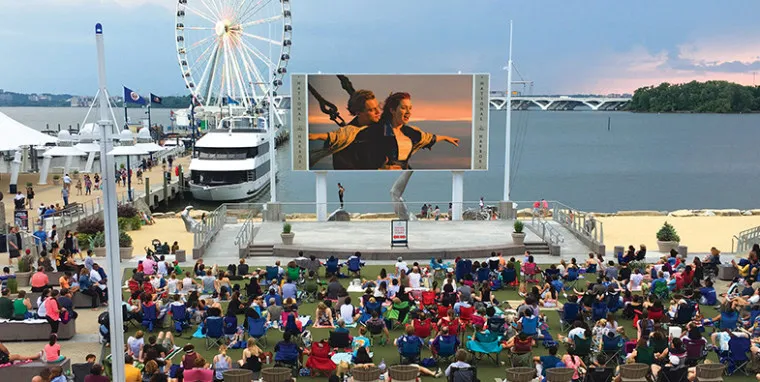 A large crowd sits outdoors near a waterfront, watching a movie on a giant screen. There’s a Ferris wheel, boats, and a sandy area in the background, with a sunset sky over the water.