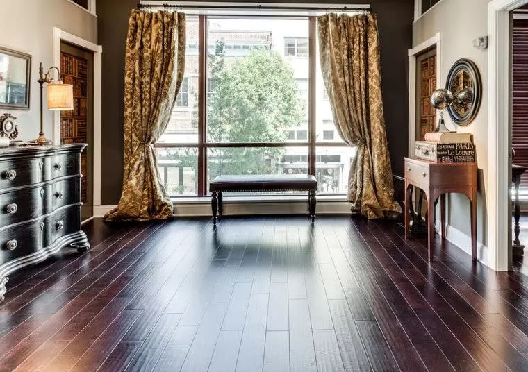 Elegant room with dark wood floor, large window with gold curtains, wooden bench, a black dresser with a lamp, and a small writing desk with books and a globe. Sunlight streams in, illuminating the space.