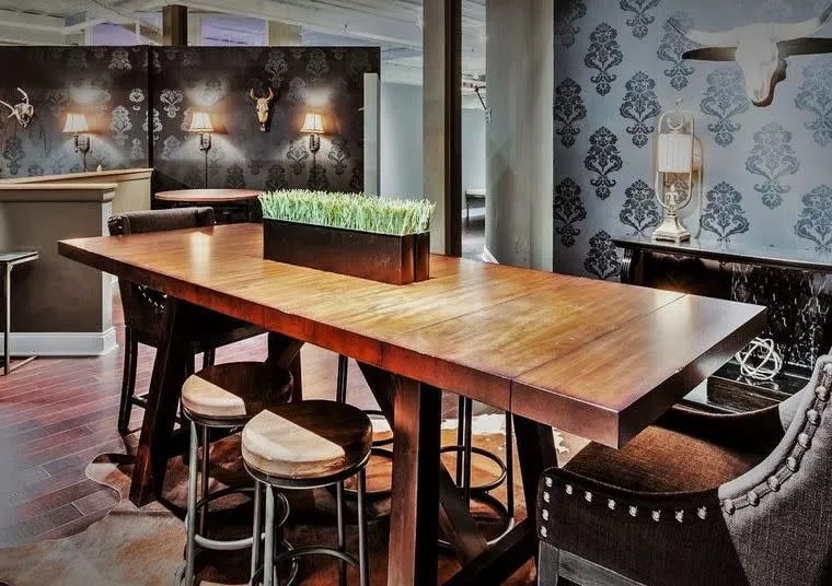 A stylish dining area with a long wooden table, round wooden stools, patterned wallpaper, wall-mounted lamps, and decorative animal skulls. A rectangular planter with grass sits at the center of the table.