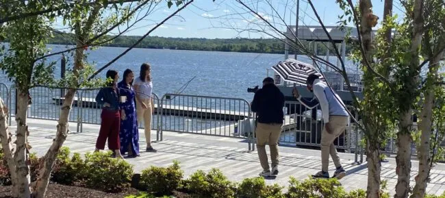 Two people pose for a photo by a waterfront while a photographer takes their picture. Another person holds an umbrella to shade them. Trees and railings frame the scene, with blue sky and water in the background.