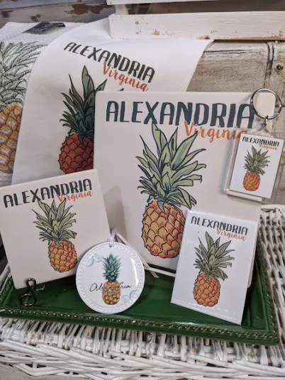 Merchandise featuring a pineapple illustration and the text Alexandria Virginia, including a sign, towel, coaster, ornament, magnet, and keychain, arranged on a green tray atop a white wicker surface.