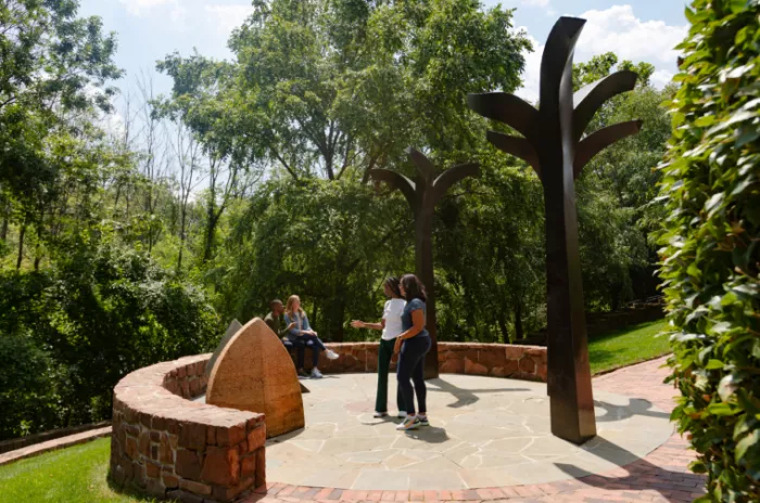 Several people gather in a circular stone plaza with tall, abstract metal sculptures resembling trees, surrounded by lush greenery and trees under a partly cloudy sky.