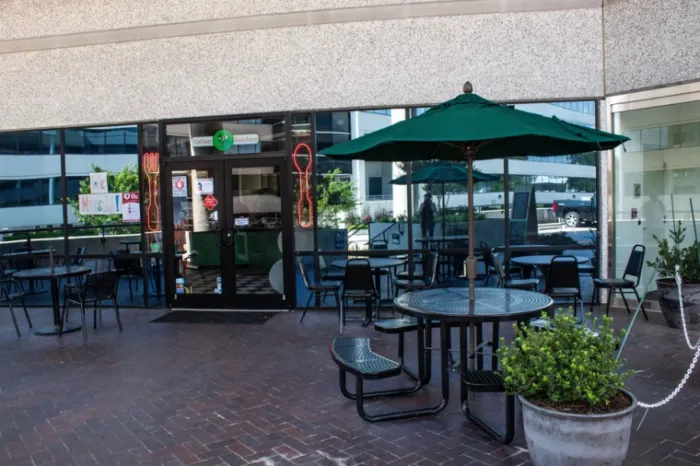 Outdoor patio with metal tables and chairs, some shaded by green umbrellas, outside a building entrance with glass doors. Potted plants and signs are visible near the door and on the brick-paved terrace.