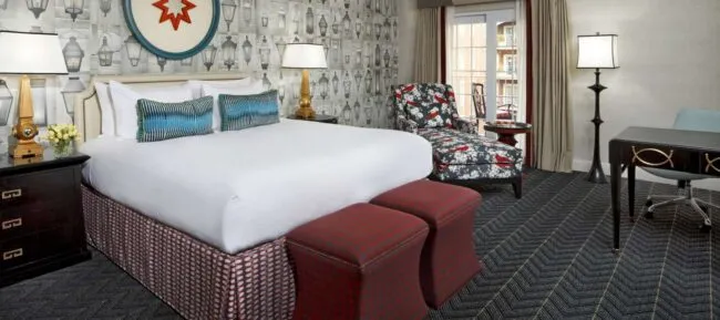 A stylish hotel room with a king bed, patterned ottomans, floral chaise lounge, desk with blue chair, large window, and wallpaper featuring black and white lantern illustrations.