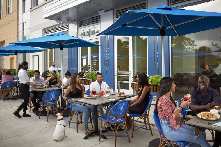 People are dining outdoors at a restaurant with blue umbrellas and chairs. Two servers are helping customers, and a dog sits beside one table. The atmosphere is lively and relaxed, with groups enjoying food and conversation.