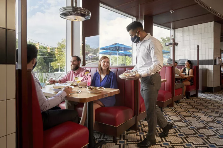A waiter wearing a mask serves food to three people seated at a booth in a diner with red seats and large windows. The group smiles and talks while enjoying their meal. Other customers are seated in the background.