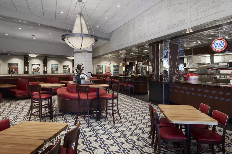 A retro-style diner with red booths, wooden tables, and red chairs. The floor has a geometric tile pattern, and a large round light fixture hangs from the ceiling. The counter and kitchen are visible in the background.