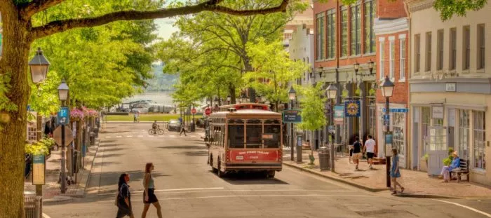 A red trolley drives down a tree-lined street with shops, pedestrians, and lampposts on either side. People walk on sidewalks and cross the street under bright daylight. In the distance, water is visible.