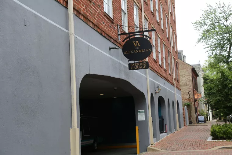 A brick building with a sign reading The Alexandrian Parking Garage above an entrance to an indoor parking area, next to a cobblestone sidewalk lined with trees.