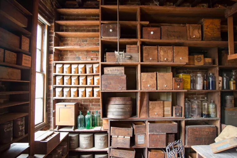 Wooden shelves and cubbies filled with old wooden boxes, jars, bottles, and tins line a brick wall in a vintage apothecary or general store, with sunlight streaming through a tall window on the left.