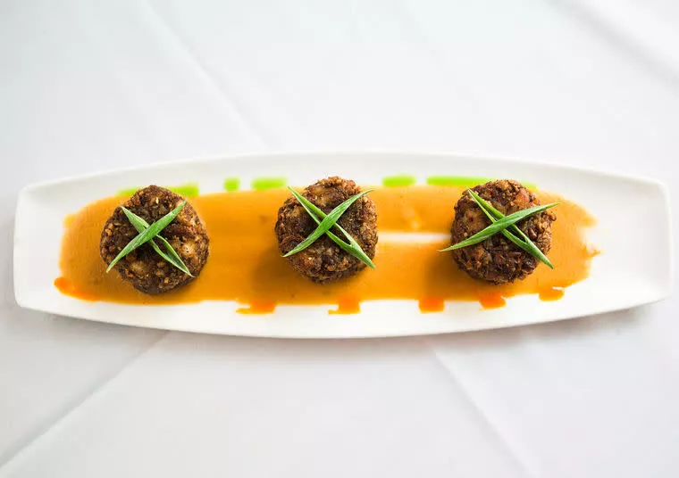 Three round crab cakes are arranged in a row on a rectangular white plate, topped with green garnish and served on a bed of orange sauce, all set on a white tablecloth.